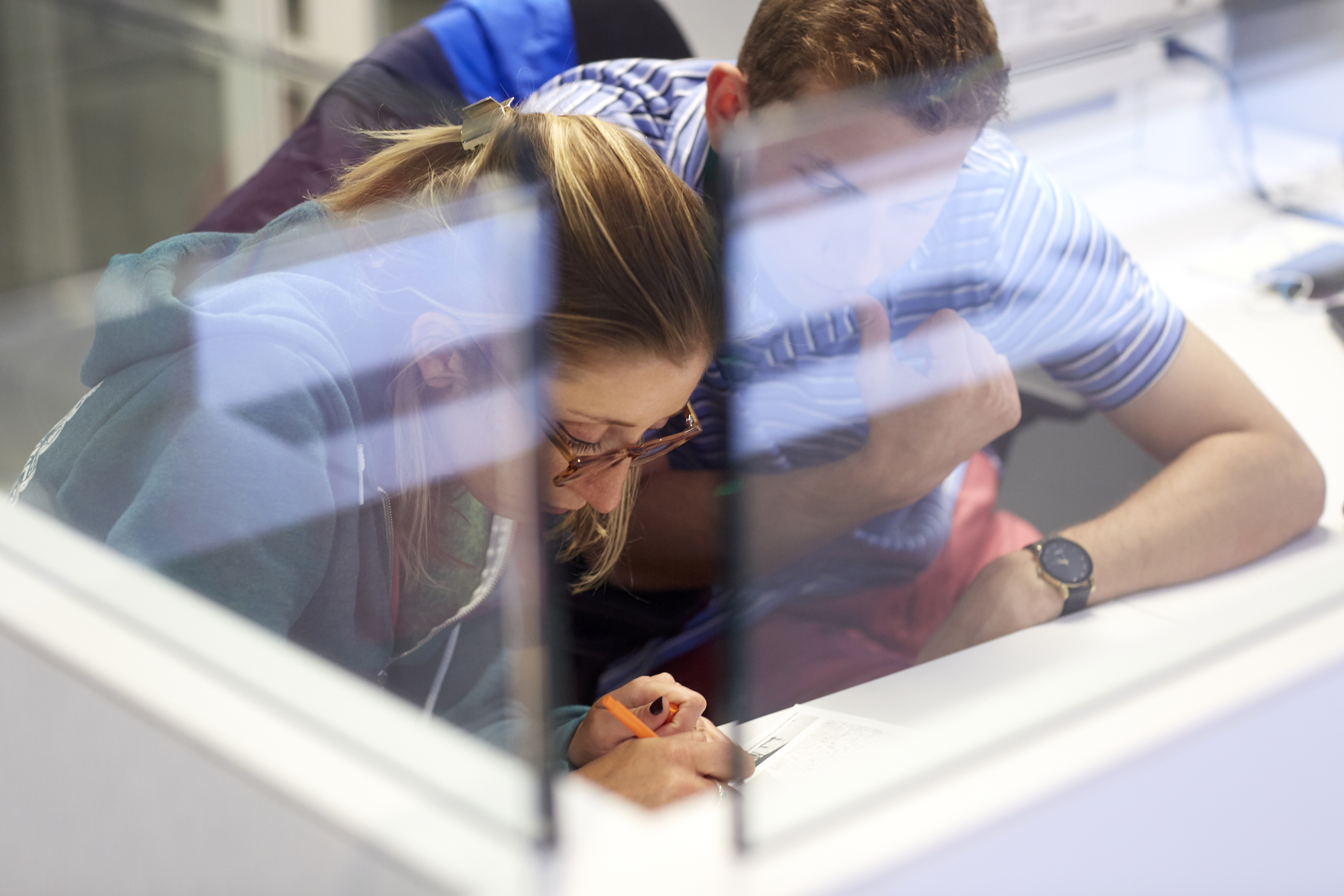 Two colleagues sit at a workstation, one writing on a piece of paper while the other observes.